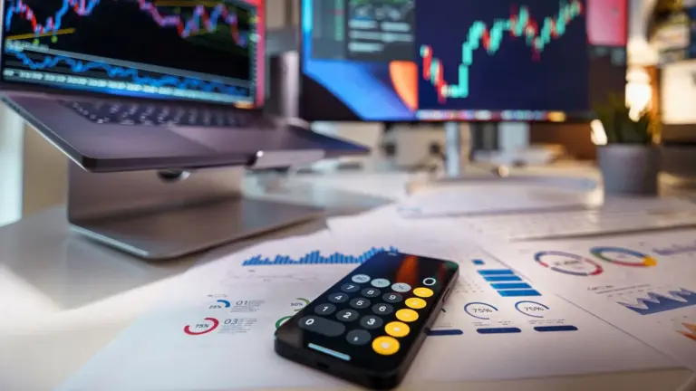Laptop and monitor showing market charts beside reports and a calculator.